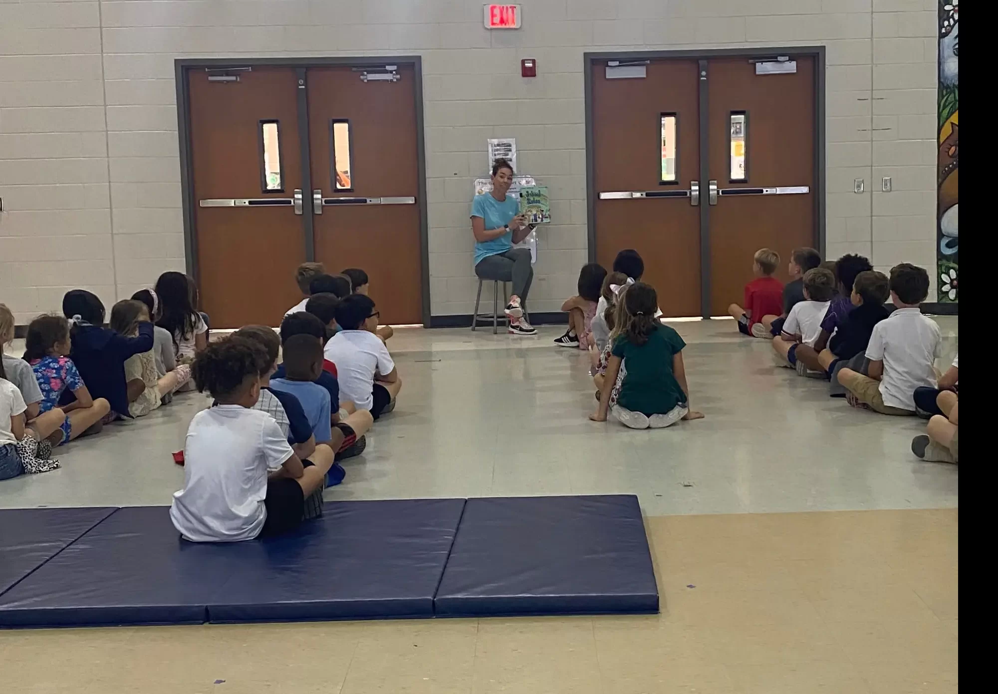 PE teacher reading School Rules book to elementary students sitting in organized rows during first day physical education lesson plan